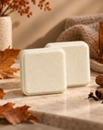 Two square off-white bath bombs with a textured finish, styled on a marble surface with autumn leaves, cinnamon sticks, a candle, and a vase in the background.