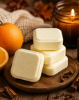 Four square cream-colored soap bars stacked on a wooden board, styled with a whole and halved orange, cinnamon sticks, cloves, mint leaves, a lit amber candle, and a small dark bottle on a rustic wooden surface.