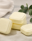 Three pale yellow square bath salts or soap bars arranged on a light stone surface with a cream cloth, green eucalyptus leaves, and lavender sprigs in the background.