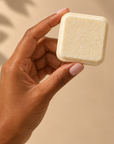 A hand with light pink manicured nails holds a small, square, off-white bar product against a warm beige background with soft leaf shadows.