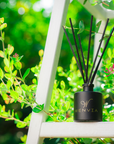 A black reed diffuser with clear branding and gold lettering, featuring multiple black reeds inserted into the bottle, placed on a wooden surface with foliage in the background.