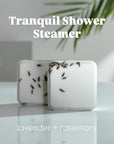 Two white shower steamers with lavender buds on a reflective surface, labeled 'Tranquil Shower Steamer' and 'lavender + rosemary' which is its aromatic scent.