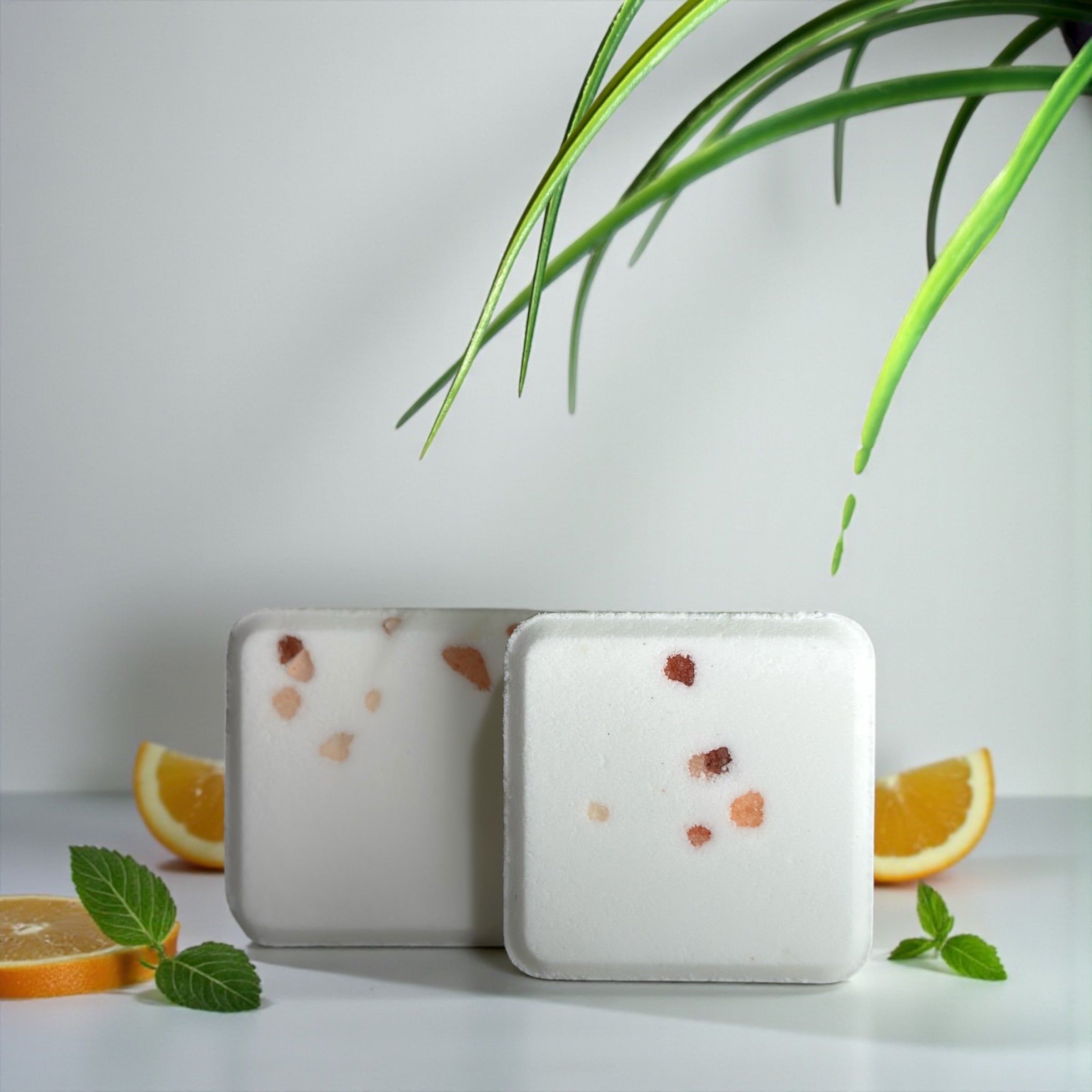 Two square shower steamers with visible ingredients on a white surface with orange slices and green leaves.