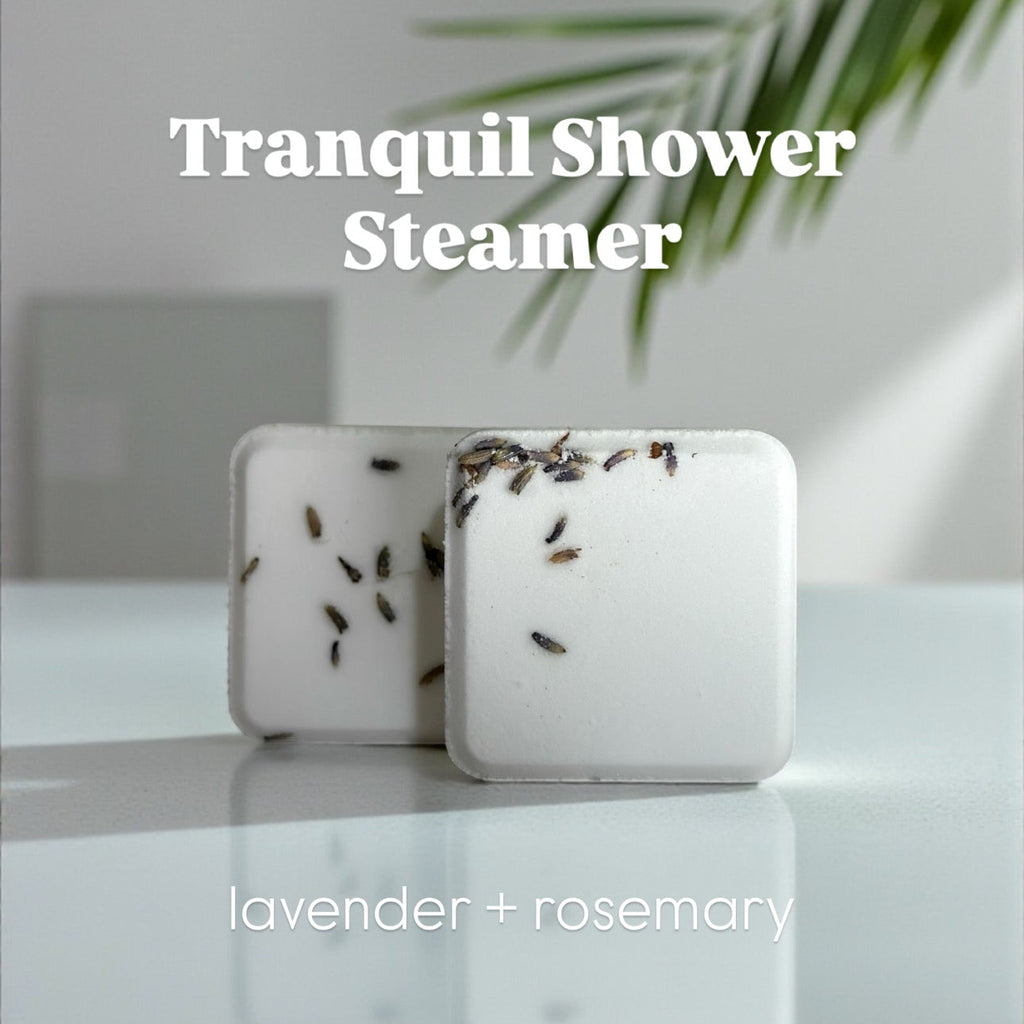 Two white shower steamers with lavender buds on a reflective surface, labeled 'Tranquil Shower Steamer' and 'lavender + rosemary' which is its aromatic scent.